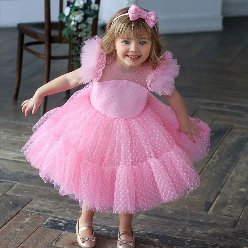 Adorable girl in the 'Ashlyn' Dotted Swiss Dress in princess pink, showcasing a whimsical full skirt and ruched tulle sleeves, ideal for parties.